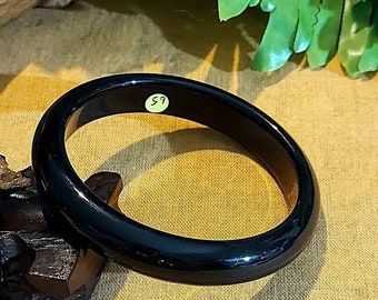 May include: A black bangle bracelet with a smooth, glossy finish. The bracelet is on a beige fabric surface. The number "59" is visible on the bracelet.