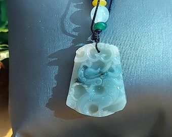 Puede incluir: Collar con colgante de jade tallado. El colgante es de color verde claro con detalles en verde oscuro. El collar tiene un cordón negro con cuentas amarillas, blancas y verdes. El colgante mide aproximadamente 5 cm de largo.