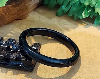 May include: A glossy, black bangle bracelet. The smooth, circular bracelet is displayed on a dark, carved wooden stand against a backdrop of green foliage and a tan surface. The bracelet is a simple, elegant piece of jewelry.