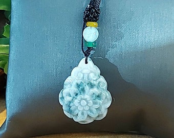 Puede incluir: Un colgante de jade verde tallado en forma de flor, colgado de un cordón negro con cuentas verdes y amarillas.