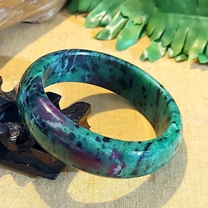 Puede incluir: Un brazalete de jade verde y rosa con un patrón moteado. El brazalete está hecho de piedra natural y tiene un acabado liso y pulido.