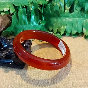 Puede incluir: Un brazalete de piedra preciosa pulida, translúcida, de color rojo anaranjado. El brazalete es liso y circular, con una etiqueta en el interior. El fondo incluye follaje verde y una superficie beige.