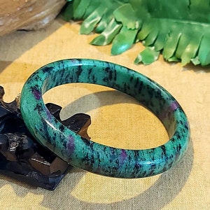 Puede incluir: Un brazalete de jade verde y morado. El brazalete está hecho de una piedra lisa y pulida y tiene un color verde oscuro con vetas moradas.
