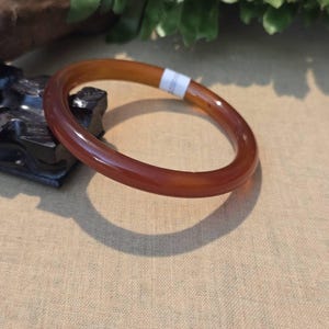 May include: A polished, reddish-brown bangle bracelet. The bracelet has a smooth, glossy finish and a circular design. The color transitions from a lighter brown to a deeper, richer red. The bracelet is resting on a neutral-toned surface.