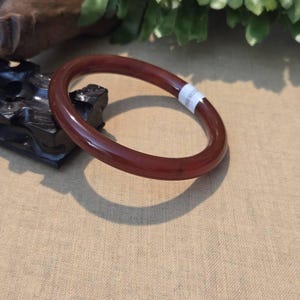 May include: A polished, reddish-brown bangle bracelet. The circular bracelet is made of a solid material and has a smooth, glossy finish. A white sticker with black text is wrapped around the bracelet. The bracelet is resting on a tan surface.