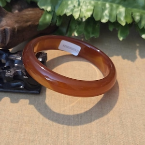 May include: A polished, amber-colored jade bangle bracelet. The smooth, circular bracelet is displayed on a neutral surface, with a tag that reads "Y2502020331". The bracelet is a classic jewelry piece.