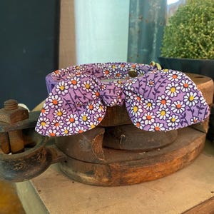 May include: A purple pet collar with a daisy pattern bow tie. The bow tie features white daisies with yellow centers on a purple background. The collar is resting on a wooden surface.