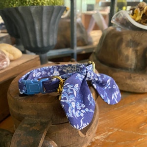 Op de afbeelding: Een blauw-witte halsband voor huisdieren met een bloemenpatroon en een stoffen strik. De halsband is voorzien van een lichtblauwe plastic gesp en een goudkleurige metalen D-ring voor het bevestigen van penningen, gemaakt van duurzame stof.