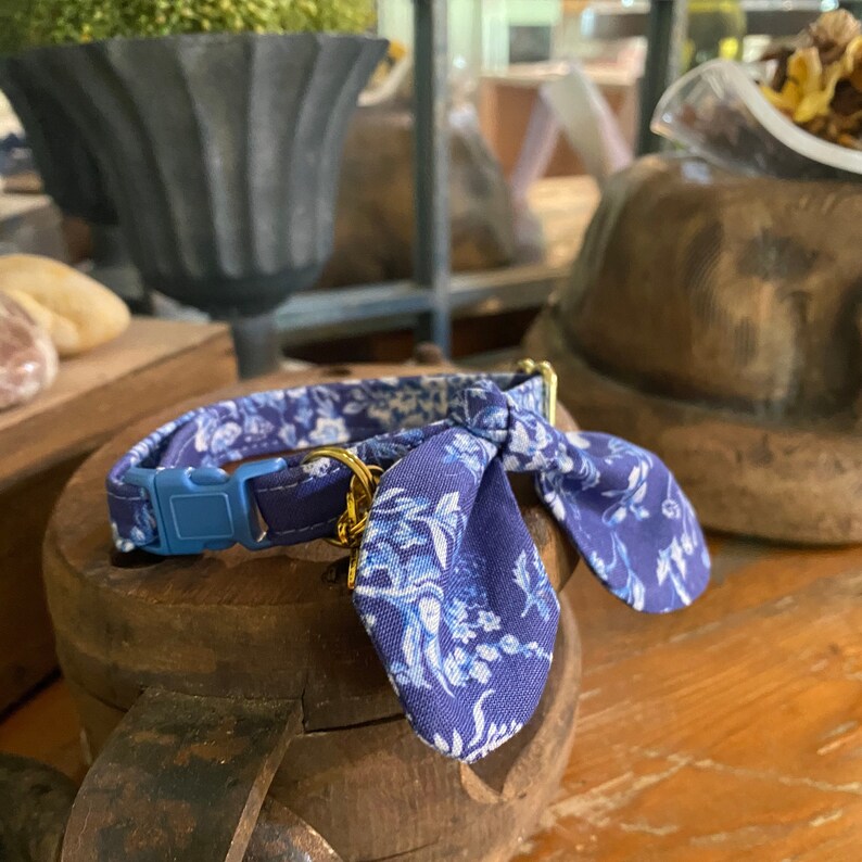 Op de afbeelding: Een blauw-witte halsband voor huisdieren met een bloemenpatroon en een stoffen strik. De halsband is voorzien van een lichtblauwe plastic gesp en een goudkleurige metalen D-ring voor het bevestigen van penningen, gemaakt van duurzame stof.