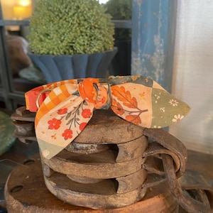 May include: A pet collar featuring a fabric bow tie with a patchwork design of orange gingham, autumn leaves, and small red and white flowers. The collar includes an orange plastic buckle and a gold-toned metal D-ring for attaching a leash.