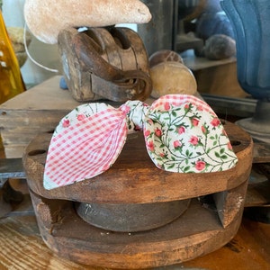 May include: A pink and white gingham bow with a floral print on the other side. The bow is tied around a wooden spool.