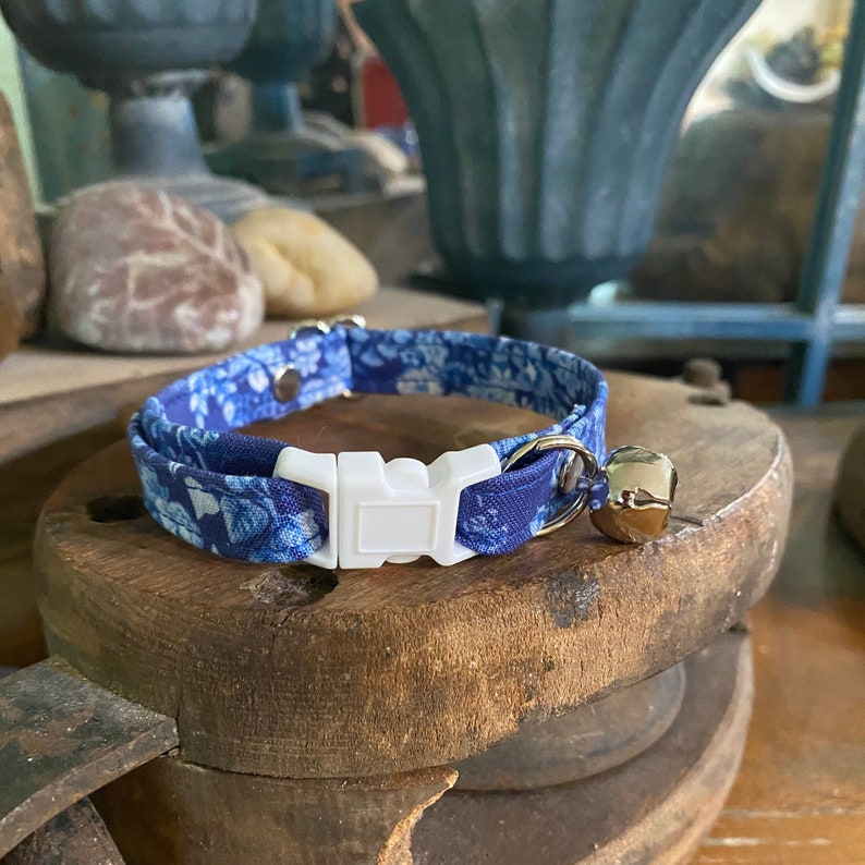 Op de afbeelding: Een blauwe stoffen halsband voor huisdieren met een bloemenpatroon in diverse blauwtinten. De halsband is voorzien van een witte plastic klikgesp, een zilverkleurige metalen D-ring en een klein, rond, glanzend zilverkleurig belletje.