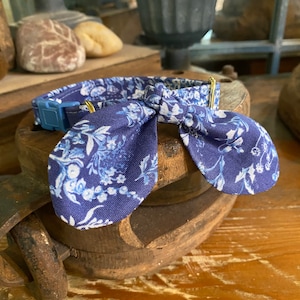 Op de afbeelding: Een blauwe stoffen halsband met een wit bloemenpatroon en een bijpassende strik. De halsband is voorzien van een blauwe plastic klikgesp en goudkleurige metalen hardware voor verstelling, met een gedetailleerd botanisch printontwerp.