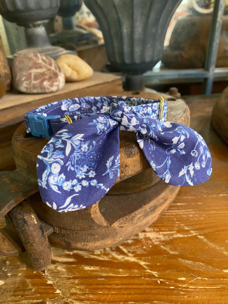 Op de afbeelding: Een blauwe stoffen halsband met een wit bloemenpatroon en een bijpassende strik. De halsband is voorzien van een blauwe plastic klikgesp en goudkleurige metalen hardware voor verstelling, met een gedetailleerd botanisch printontwerp.
