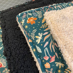 Pu&ograve; includere: Una pila di coperte piegate caratterizzate da un tessuto verde ottanio scuro con un motivo ripetuto di fiori arancioni, funghi rosa e falene gialle. Le coperte sono foderate con spesso pile sherpa testurizzato nei colori nero e bianco crema.