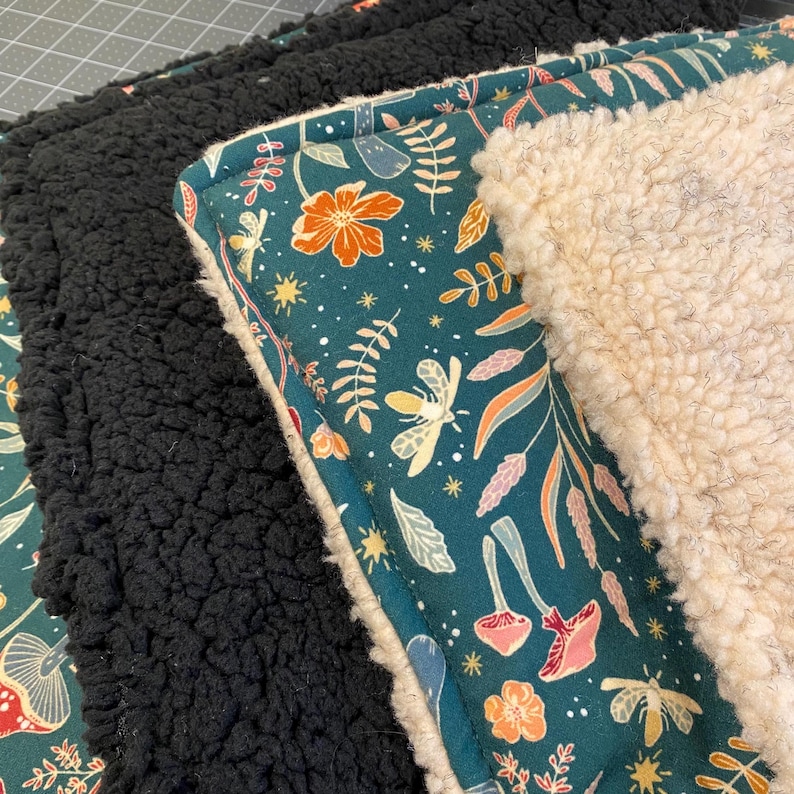 Pu&ograve; includere: Una pila di coperte piegate caratterizzate da un tessuto verde ottanio scuro con un motivo ripetuto di fiori arancioni, funghi rosa e falene gialle. Le coperte sono foderate con spesso pile sherpa testurizzato nei colori nero e bianco crema.