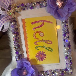 May include: A rectangular picture frame decorated with purple beads, rhinestones, and two large purple fabric flowers. A gold bow charm is attached to the top left corner. The frame contains a yellow card with the word hello and a flower graphic.