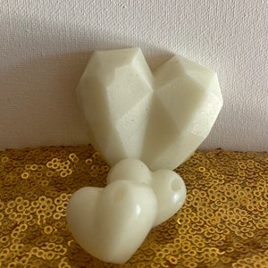 May include: Ivory-colored heart-shaped wax melts. One large heart has a geometric faceted design, while two smaller hearts have a smooth, rounded shape. The melts are on a gold sequined surface.