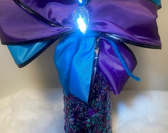 Puede incluir: Una botella decorativa con un lazo morado y turquesa. La botella está cubierta de purpurina y lentejuelas en forma de estrella. Un lazo grande, morado con ribete negro y un acento turquesa se encuentra en la parte superior de la botella. Una gema grande y transparente está en el centro del lazo.