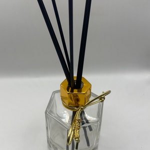 May include: A clear glass diffuser bottle with a gold-colored hexagonal top and decorative gold accents. Black reeds are inserted into the bottle. The diffuser is designed for home fragrance.