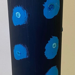 Puede incluir: Un jarrón cilíndrico alto pintado de azul marino con formas irregulares en forma de flor de color azul claro. Cada forma tiene una pequeña perla iridiscente en el centro. El jarrón es un objeto decorativo. Altura aproximada 20 cm.