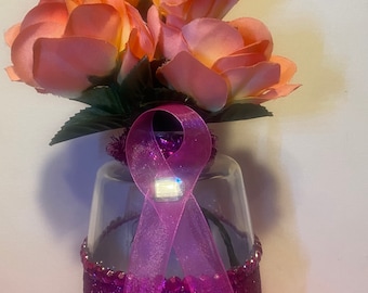 Puede incluir: Un jarrón de cristal decorativo con una cinta rosa y un ramo de rosas color melocotón. El jarrón está adornado con purpurina rosa y pedrería. El tul rosa añade un acento suave y fluido.