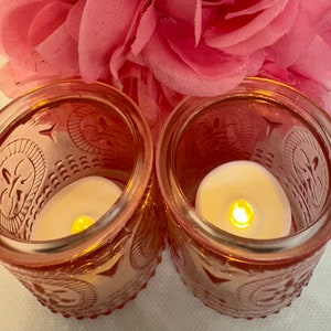 May include: Two pink glass votive candle holders with decorative embossed patterns. Each holder contains a small, glowing, battery-operated candle. A pink fabric flower is in the background.