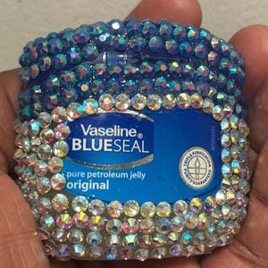Puede incluir: Un envase azul de vaselina Blue Seal, adornado con pedrería iridiscente. La etiqueta del envase es visible, con las palabras "Vaseline BLUE SEAL pure petroleum jelly original". La pedrería está dispuesta alrededor de los bordes.