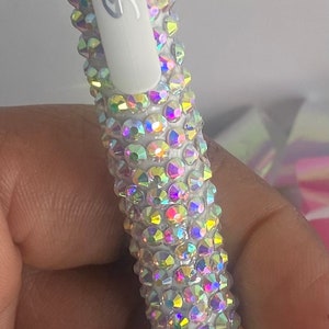 May include: A white pen with a 0.7 tip size, adorned with iridescent rhinestones. The pen has a silver tip and a white, textured grip. The pen's body is covered in colourful, sparkling crystals.