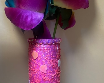 Puede incluir: Un jarrón decorativo con rosas arcoíris. El jarrón cilíndrico rosa brillante está adornado con purpurina y detalles de conchas marinas. Las rosas tienen pétalos amarillos, azules y morados.