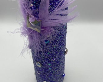 Puede incluir: Un vaso cilíndrico cubierto de purpurina en tonos azul oscuro y morado. El vaso está adornado con plumas de lavanda, gemas iridiscentes y un pequeño adorno cuadrado. El diseño general es decorativo y llamativo.