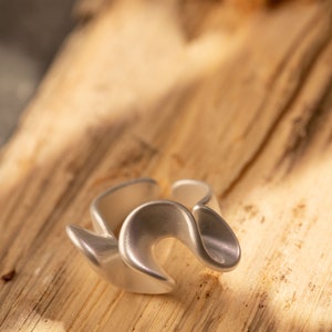 May include: A silver ring with a unique, abstract design. The ring is made of a smooth, shiny metal and has a wavy, organic shape.