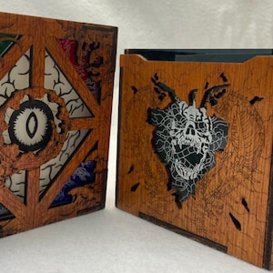 May include: Two wooden boxes with intricate laser-cut designs. The box on the left features a circular eye design with four surrounding panels, each with a different colored design. The box on the right features a skull design with a dragon-like creature surrounding it. Both boxes have a wood grain finish.