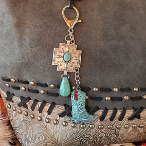 May include: A silver charm with a turquoise stone and a silver cross charm attached to a silver chain. The charms are hanging from a silver key ring. The key ring is attached to a gray leather bag with black stitching and silver studs.