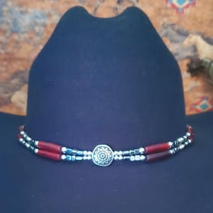 May include: A dark navy blue felt hat featuring a double-strand hat band. The band consists of cylindrical red beads, small metallic silver spheres, and dark metallic square beads, centred with a round silver medallion with a sunburst design.
