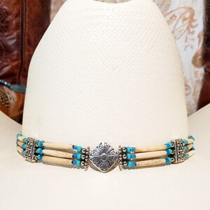 May include: A white cowboy hat with a turquoise and bone bead hat band. The hat band has a silver metal clasp with a decorative design.