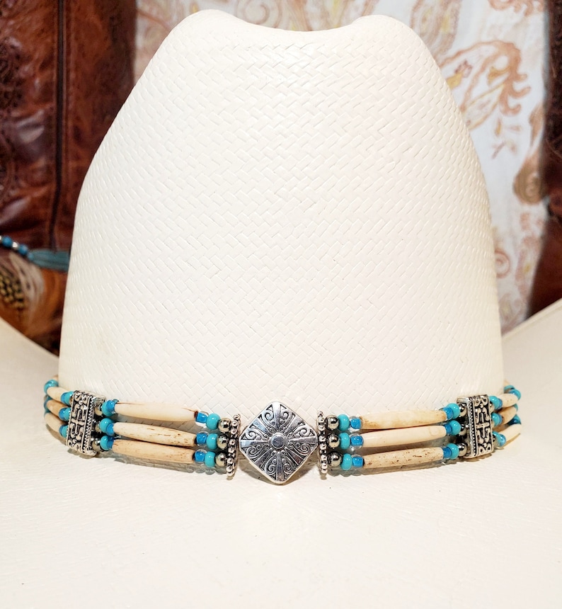 May include: A white cowboy hat with a turquoise and bone bead hat band. The hat band has a silver metal clasp with a decorative design.