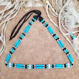 May include: A turquoise and black beaded necklace with silver accents and a leather cord. The necklace is laid out on a brown surface.