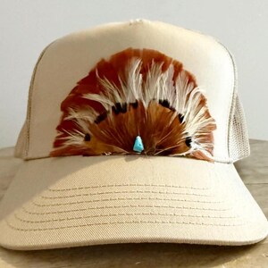 Puede incluir: Gorra de camionero color beige con un dise&ntilde;o de plumas marrones y blancas en la parte delantera. Las plumas est&aacute;n acentuadas con una peque&ntilde;a piedra turquesa. La gorra tiene una parte trasera de malla y una visera curva.