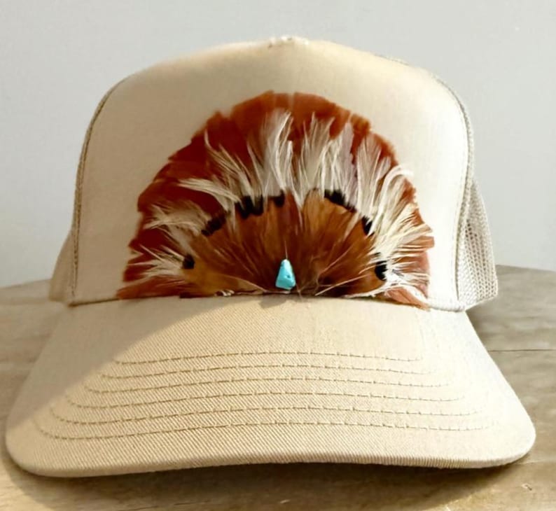 Puede incluir: Gorra de camionero color beige con un dise&ntilde;o de plumas marrones y blancas en la parte delantera. Las plumas est&aacute;n acentuadas con una peque&ntilde;a piedra turquesa. La gorra tiene una parte trasera de malla y una visera curva.