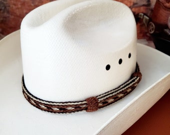Op de afbeelding: Een witte cowboyhoed met een bruin en wit gevlochten hoedband. De hoed heeft drie zwarte stippen op de kroon.
