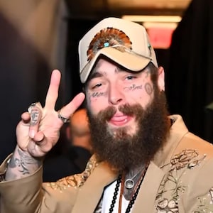 Puede incluir: Gorra de b&eacute;isbol beige con plumas y detalles turquesas. La persona lleva una blazer beige con bordados florales. La persona tiene tatuajes faciales y una larga barba. La persona hace el signo de la paz.