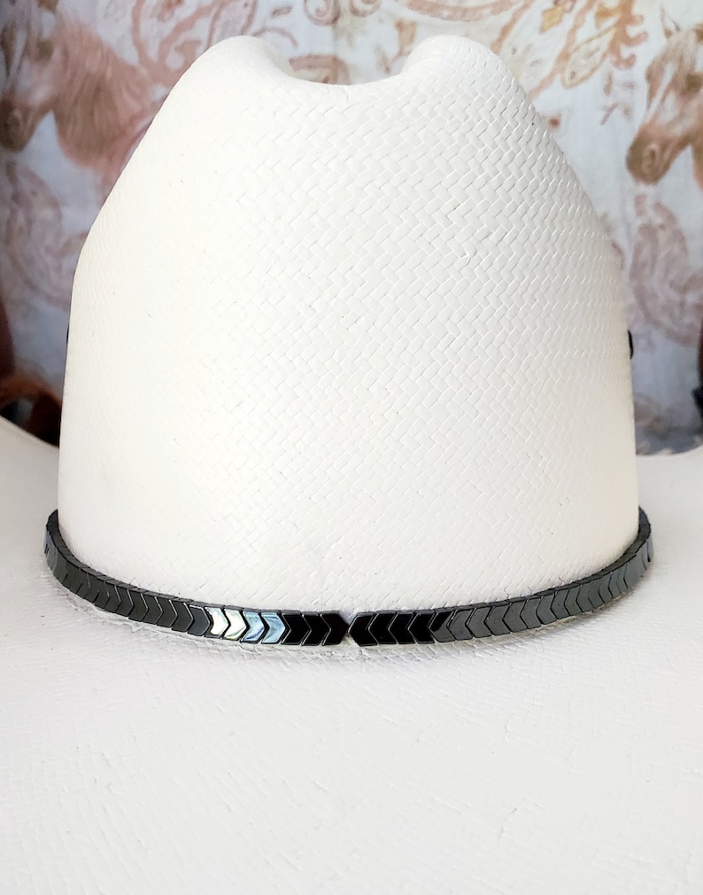 Pu&ograve; includere: Un cappello da cowboy bianco con una fascia nera a forma di freccia.