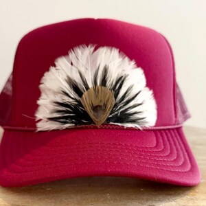 Puede incluir: Gorra de camionero burdeos con parte trasera de malla y visera curva. La gorra presenta un abanico decorativo de plumas blancas, negras y marrones en la parte delantera, a&ntilde;adiendo un toque &uacute;nico al accesorio.