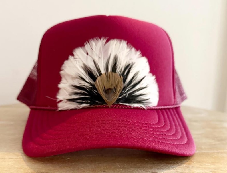 Puede incluir: Gorra de camionero burdeos con parte trasera de malla y visera curva. La gorra presenta un abanico decorativo de plumas blancas, negras y marrones en la parte delantera, a&ntilde;adiendo un toque &uacute;nico al accesorio.