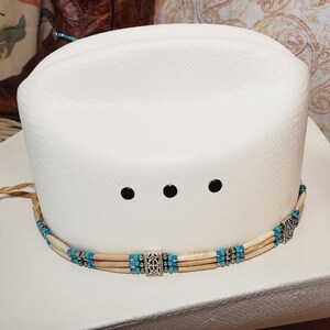 May include: White cowboy hat with a turquoise and bone bead hat band. The hat band has three silver accents.