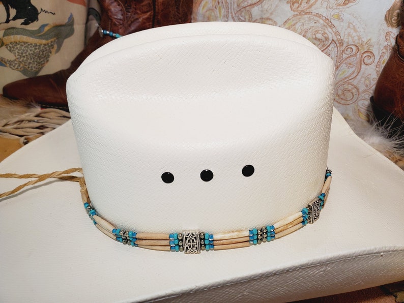 May include: White cowboy hat with a turquoise and bone bead hat band. The hat band has three silver accents.