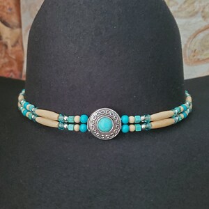 May include: A black cowboy hat with a decorative hat band. The hat band features turquoise and cream-coloured beads, with a silver-coloured circular centrepiece with a turquoise stone.