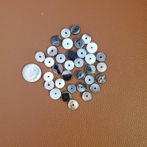 Könnte beinhalten: Eine Sammlung runder, flacher, irisierender Muschelknäpfe in den Farben Weiß, Schwarz und Grau. Die Knöpfe sind in einer lockeren Gruppe auf einer braunen Oberfläche angeordnet.