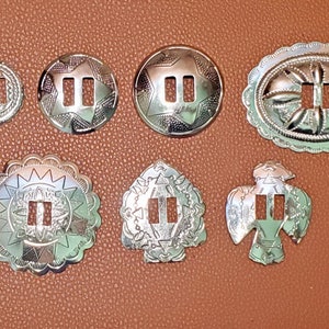 May include: Seven silver-toned metal conchos with intricate designs. Each concho has two slots for a strap or buckle. The conchos are arranged in a row on a brown leather surface.